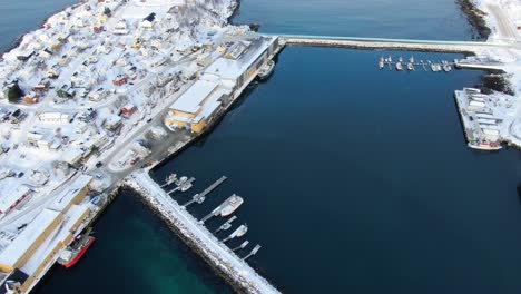 Drone-view-on-the-Tromso-mountains-in-winter-full-of-snow-showing-Husoy-a-small-town-on-an-island-surrounded-by-the-sea-and-its-small-port-with-flying-seagulls