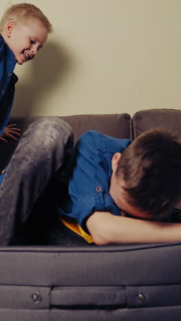 two brothers are playing on the couch with travelling bag in the living-room. elder boy is trying to lay himself into a grey suitcase at home. vertical video