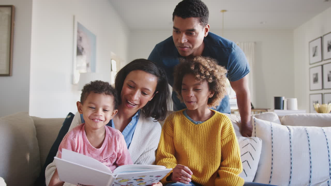 Giovane mamma afroamericana che legge un libro con i suoi due figli, il papà che guarda da dietro le loro spalle.