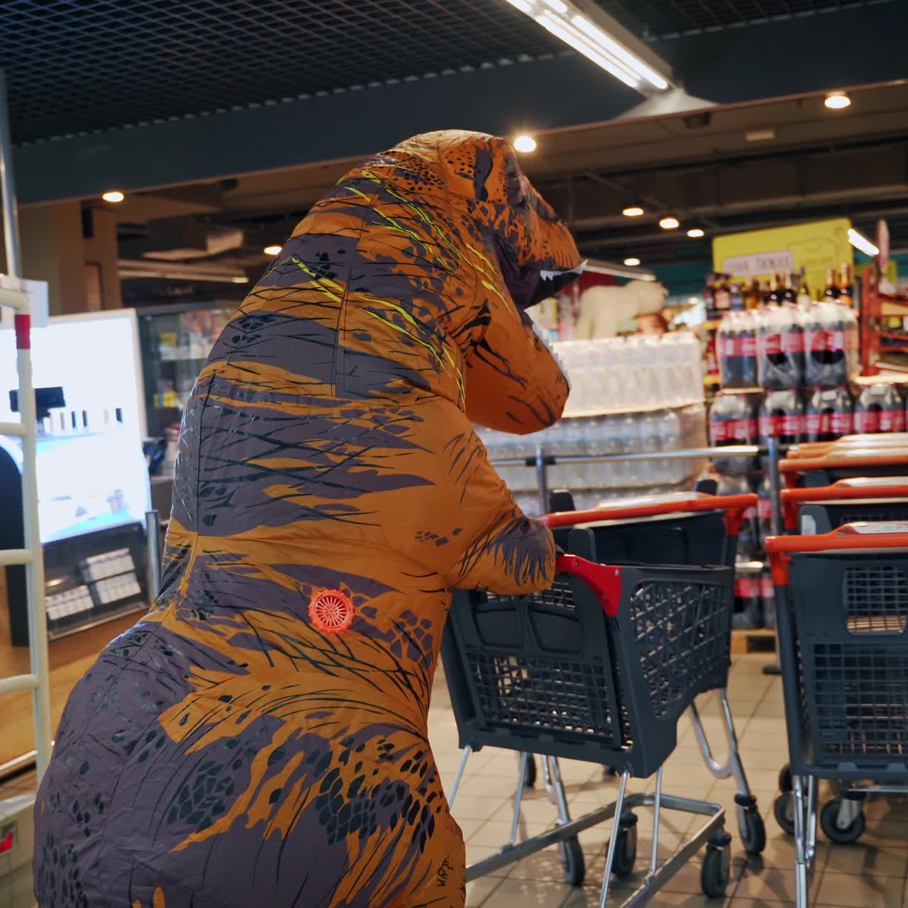 Høj, orange dinosaur, der går forbi supermarkedet og skubber en tom indkøbsvogn. Sjov T-Rex passerer kasserne og efterlader vognen