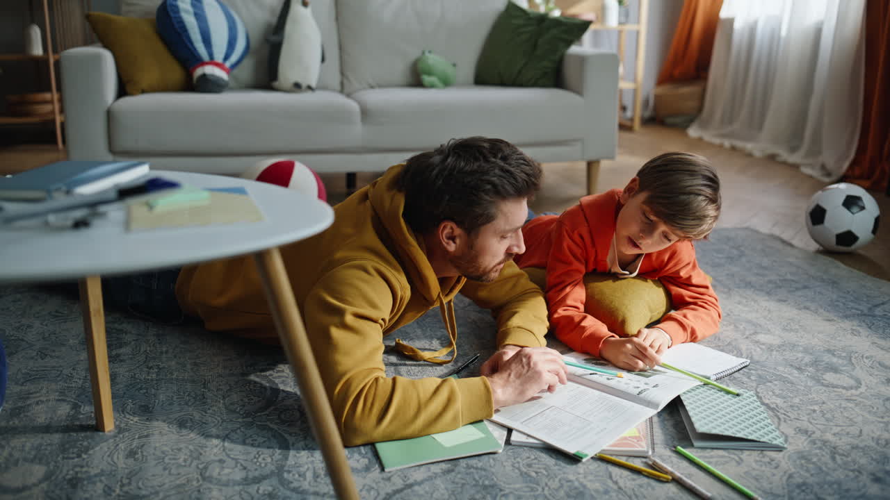 Primer plano de un padre apoyando a su hijo estudiando en casa, leyendo un libro de texto juntos tumbados en el suelo.