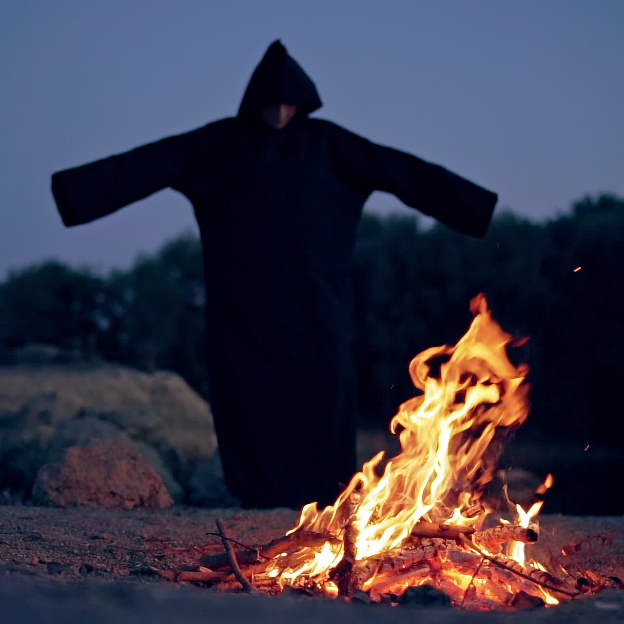 Scary view of a evil in black costume at the fire. Silhouette of death with stretched arms outdoors. Halloween evening.