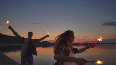 Jóvenes-Amantes,-Hombres-Y-Mujeres,-Corren-Alegremente-Por-La-Playa-Con-Podredumbre-Brillante-Y-Caliente-En-Sus-Manos-En-Cámara-Lenta.-Relación-Romántica.-Las-Bengalas-Arden-En-Las-Manos-De-Los-Amantes.