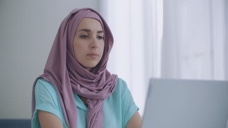 muslim woman working on a laptop in the office with her christian coworkers. closeup portrait of young muslim businesswoman in hijab using the laptop indoors in the apartment