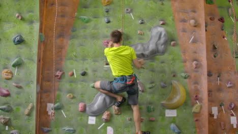 Male-climber-climbs-the-cliff-wall-in-the-room-reaching-and-gripping-hold.-A-professional-concentrated-climber-moves-up-the-wall-the-camera-rises-and-lays-down-his-movements.-Boom-and-truck-follow-camera.