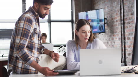 Young-Caucasian-woman-and-Asian-man-collaborate-on-a-laptop-in-a-business-office