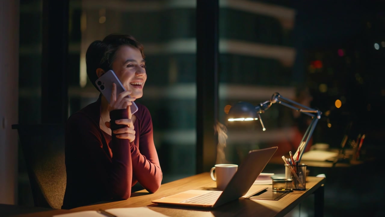 Donna d'affari sorridente al telefono in ufficio di sera. Una donna felice che lavora al laptop di notte.