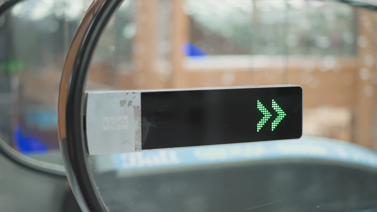 zoomed out side view of glass mounted green led arrow display pointing down at entrance of moving walkway with blurred mall decor reflected on glass under soft ambient lighting and subtle contrast