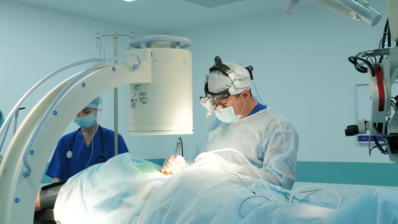 외과 의사는 수술을 수행하고 이동 현미경을 통해 볼 수 있습니다.