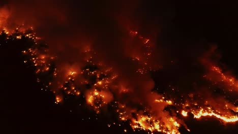 Aerial-view-of-a-raging-and-glowing-wildfire-spreading-across-woodlands,-nighttime