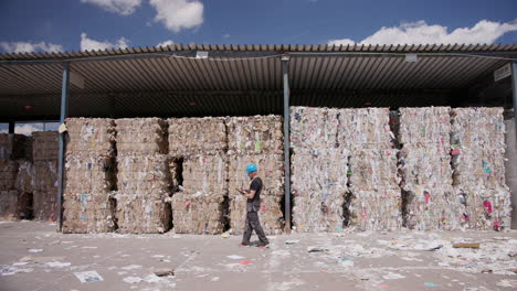 Laborer-counts-stacked-bales-of-waste-paper-stored-under-shelter,-tracking-shot