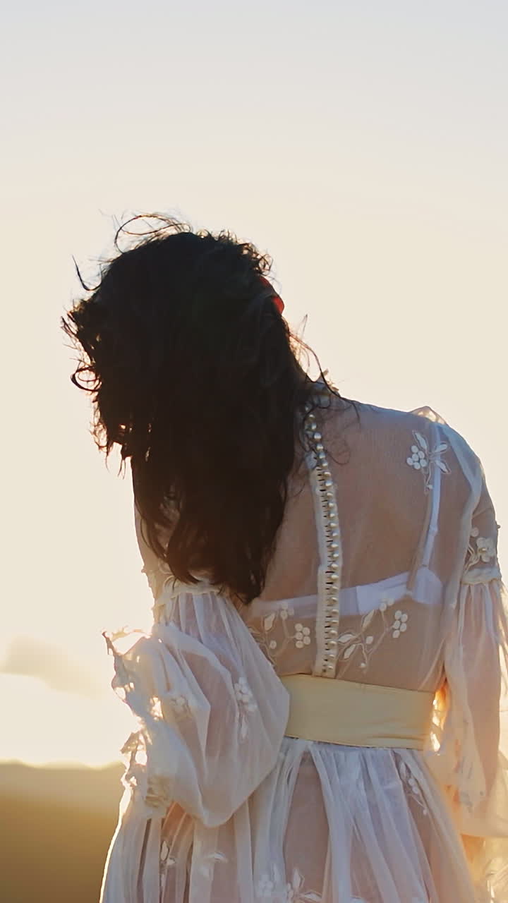 Dark-haired woman in transparent white dress standing high in the mountain. Unrecognizable lady stands her back to the camera enjoying setting sun. Vertical video