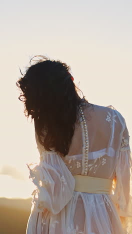 dark-haired woman in transparent white dress standing high in the mountain. unrecognizable lady stands her back to the camera enjoying setting sun. vertical video