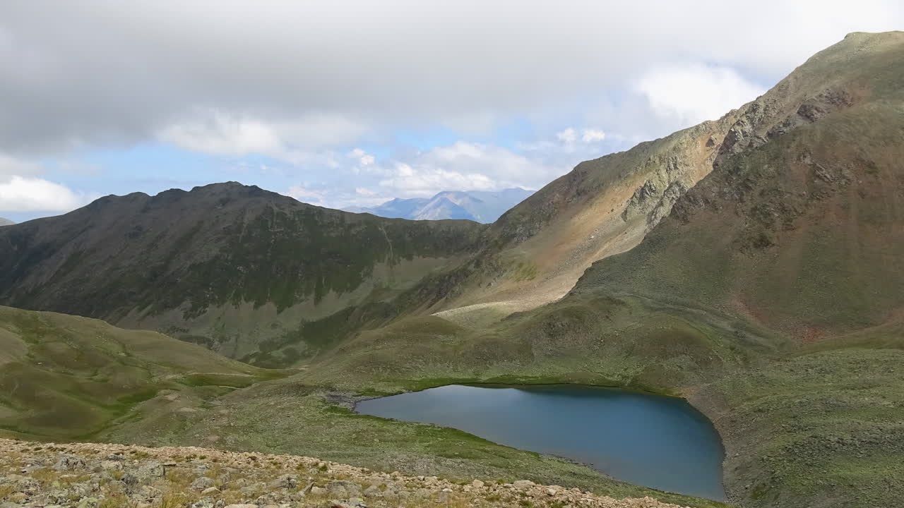 Kafkas Dağları, Rusya'da, engebeli dağ sırtları ve yeşil vadi yamaçları ile çevrili sakin bir alp gölünün hızlandırılmış görünümü, sürüklenen bulutlar ve değişen ışıkla.