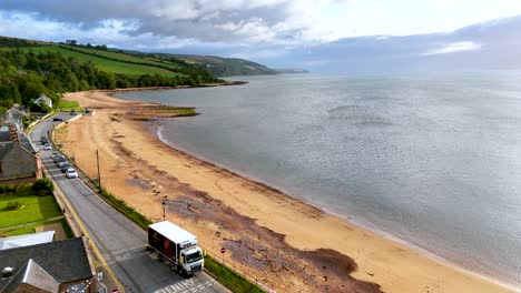 Drone camera glides above a quiet sandy beach and coastal road, revealing scenic shoreline, calm sea, and lush green hills under soft daylight