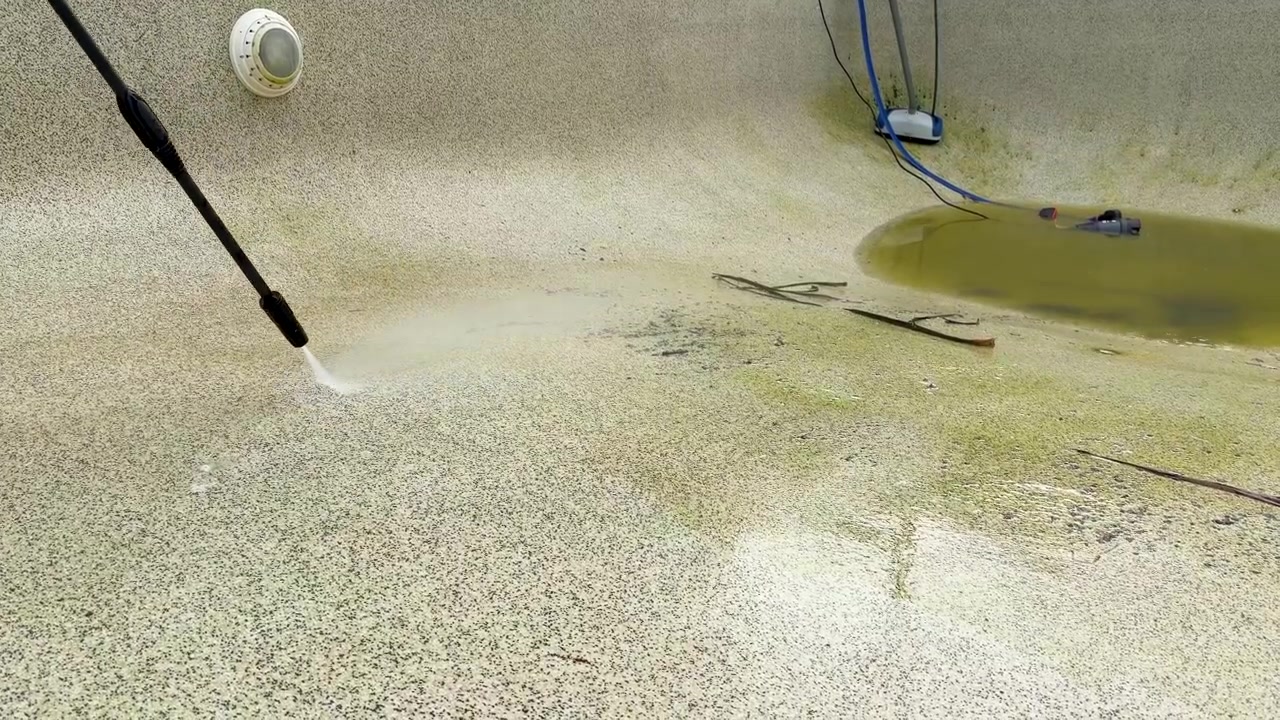 A pressure washer wand sprays water to clean algae and grime from a concrete pool.