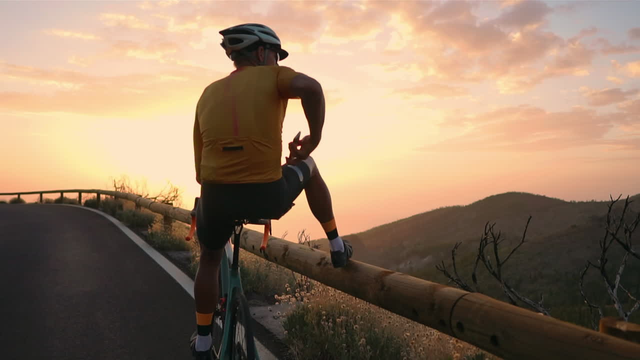 Captured in slow motion, a pro cyclist, donning a helmet and sports attire, gazes at a picturesque vista from the mountain's edge using a mobile phone