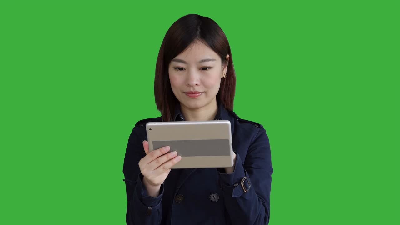푸른 코트를 입은 젊은 아시아 여성, 스마트폰, 디지털 태블릿을 사용하고 카메라를 보고, 크로마 키를 위해 녹색 화면 앞에 서있는 여성.
