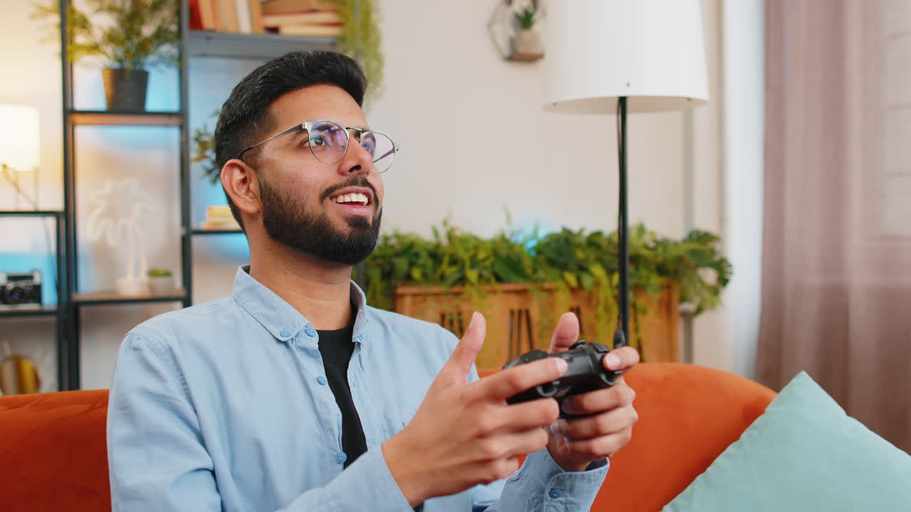Indian young man using joystick controller playing video console game online game on home sofa