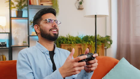 joven indio usando un controlador de joystick jugando un juego de consola de video juego en línea en el sofá de casa