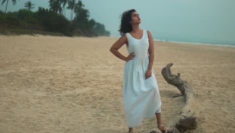 woman in white dress posing thoughtfully on a deserted beach at dusk, overcast sky