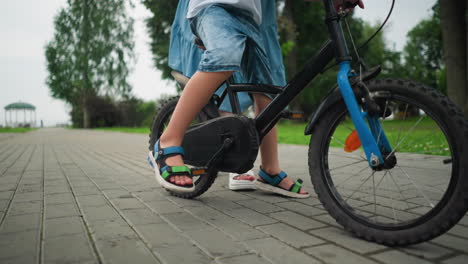 leg view of a child riding a bicycle, dropping his leg to the ground for balance, while a woman follows closely from behind