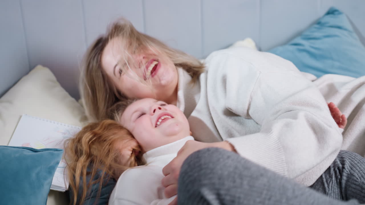 Madre sorridente e figlia condividono un gioioso momento di unione sul letto, ridendo e abbracciandosi con energia giocosa, esprimendo calore, amore, connessione e felicità familiare in un ambiente domestico accogliente