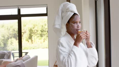 mujer biracial relajada con bata de baño y máscara en la cara bebiendo una taza de té en la ventana, en cámara lenta