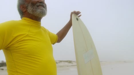 front view of thoughtful active senior african american man with surfboard standing on the beach 4k