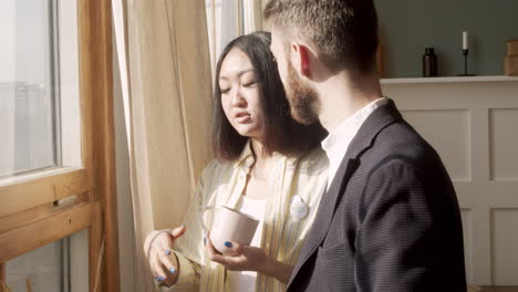 hombre con ropa formal y mujer joven sosteniendo tazas y hablando juntos mientras está de pie cerca de la ventana por la mañana