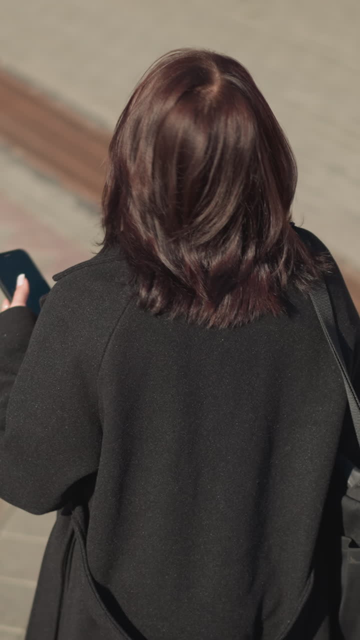 전화기에 집중하면서 머리카락을 만지고 바닥에 그늘을 던져 주위를 둘러보는 여성의 뒷면.