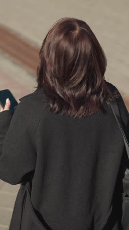 전화기에 집중하면서 머리카락을 만지고 바닥에 그늘을 던져 주위를 둘러보는 여성의 뒷면.