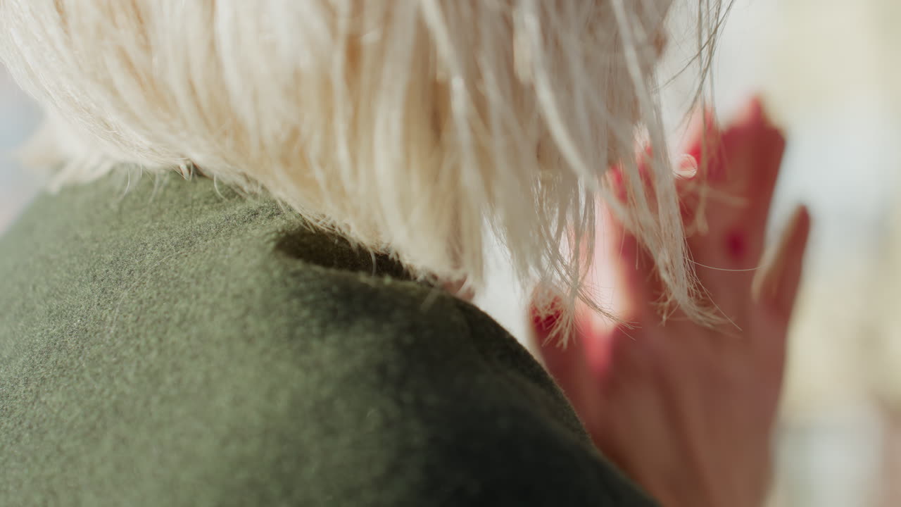 Close-up view of blonde woman outdoors with short hair illuminated by sunlight, wearing green coat, strands gently moving in breeze, capturing texture,quiet elegance and reflective atmosphere