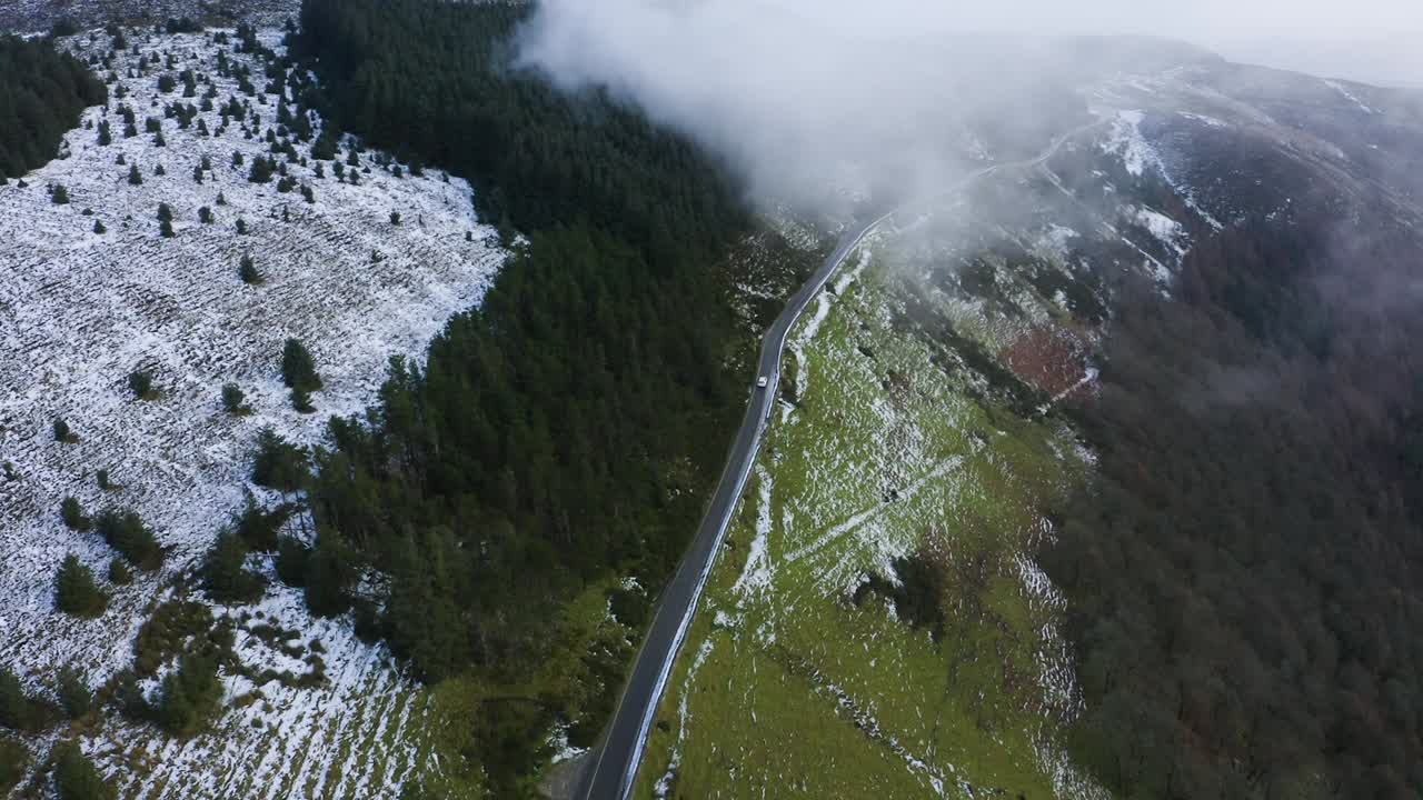 viaje a través de wicklow: seguimiento aéreo de un coche blanco en medio de montañas brumosas y bosques cubiertos de nieve conduciendo hacia las nubes