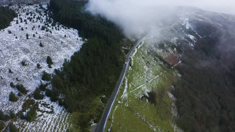 journey through wicklow: aerial tracking of a white car amidst misty mountains and snow-covered forests driving into the clouds