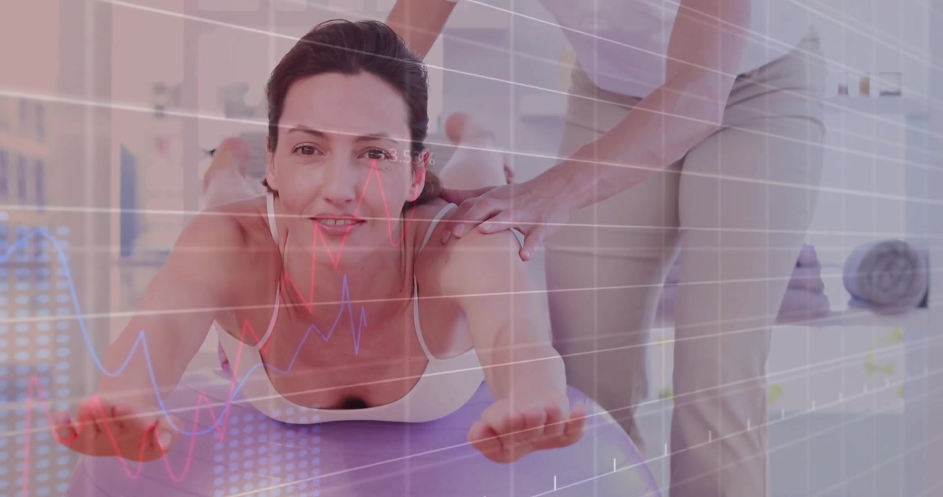 Eğitmen, fitness dersi sırasında mor top üzerinde sırt esnetme hareketini yaparken kadının omuzlarına ellerini koyarak ona rehberlik ediyor.