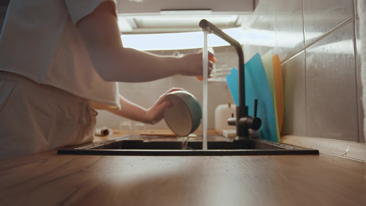 Mains rinçant un bol en céramique sous l'eau courante, nettoyage après la cuisine entre les tâches, lumière du jour douce, planches à découper colorées en arrière-plan, routine lente et intentionnelle, atmosphère de cuisine domestique rangée.