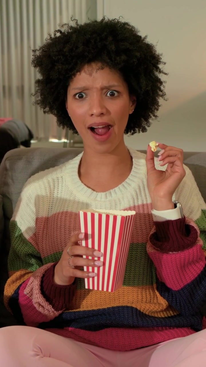 Vertikal video: Kvinde i stribet sweater spiser og reagerer på ven på sofaen, griber popcornbøtte.