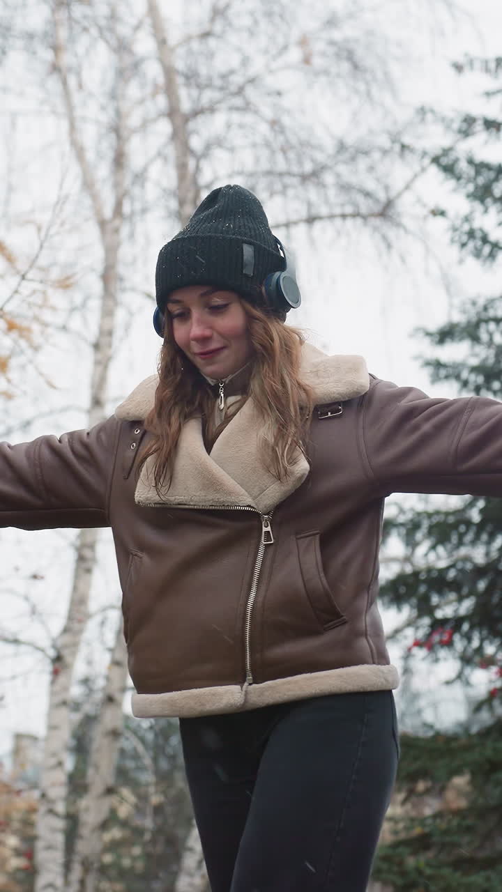 Close shot of smiling girl wearing headphone, black knit cap, brown shearling jacket, black trousers balancing gracefully with arms outstretched while walking joyfully on wide stone surface