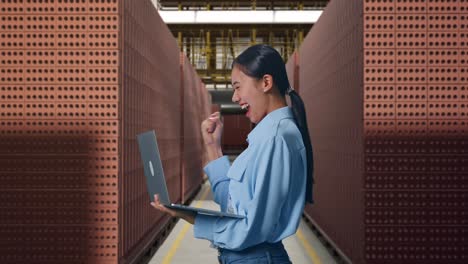side view of asian female with her laptop with red brick packed in stacks are stored, she raises her fist up with screaming goal after typed on the laptop