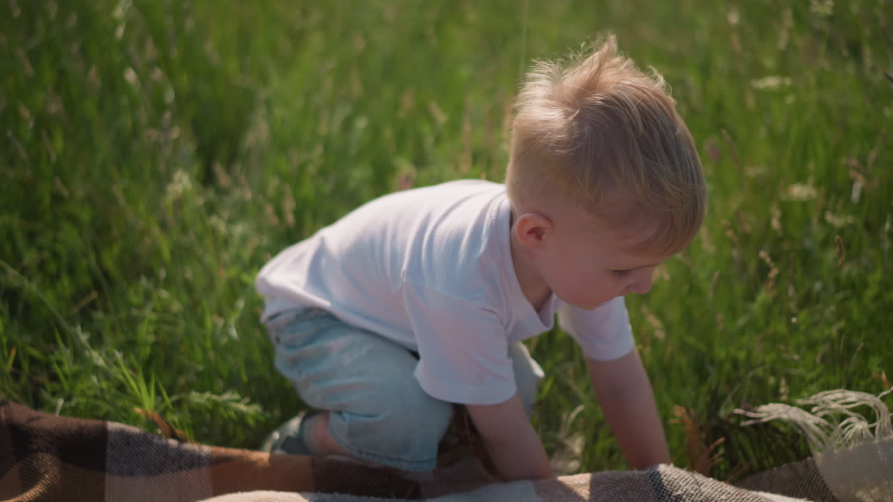 Two young children, both wearing white shirts, are seen in a sunny grass field as they lay a plaid scarf on the grass. The children are engaged in playful activity, enjoying the warmth of the day