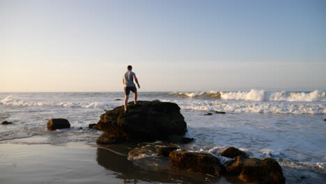 A-man-runs-in-slow-motion-on-the-beach-and-jumps-onto-a-large-rock-to-watch-the-ocean-waves-crash-on-the-coast-at-sunrise-in-Santa-Barbara,-California