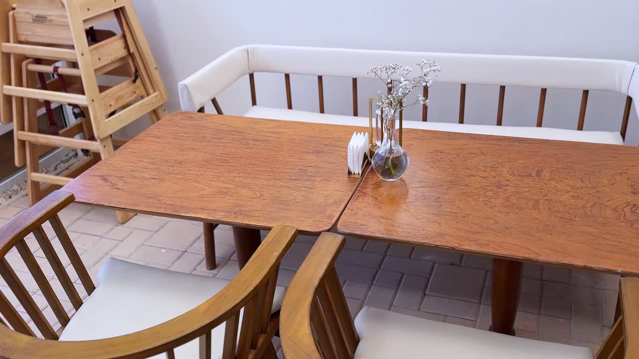 A static shot of a modern wooden table and chairs on an outdoor restaurant patio. A small vase with baby's breath flowers sits on the table. No people, quiet morning scene
