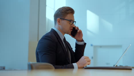 cheerful businessman talking on phone while looking at laptop monitor sitting by the desk