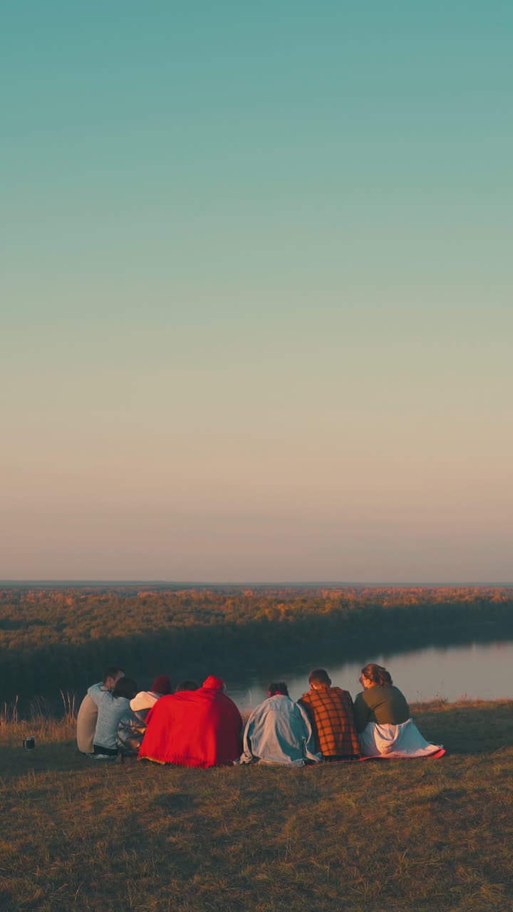 young tourists group spends time at camping with burning bonfire near tent on steep river bank in autumn evening