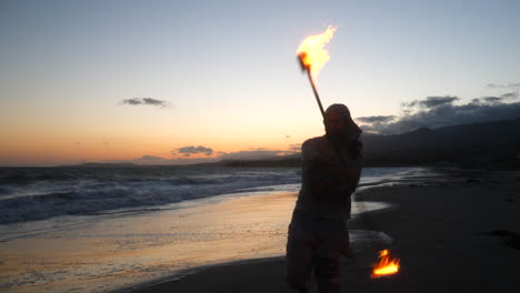 a male fire dancer or performer in silhouette spinning a flaming staff on the beach at sunset with ocean waves in slow motion