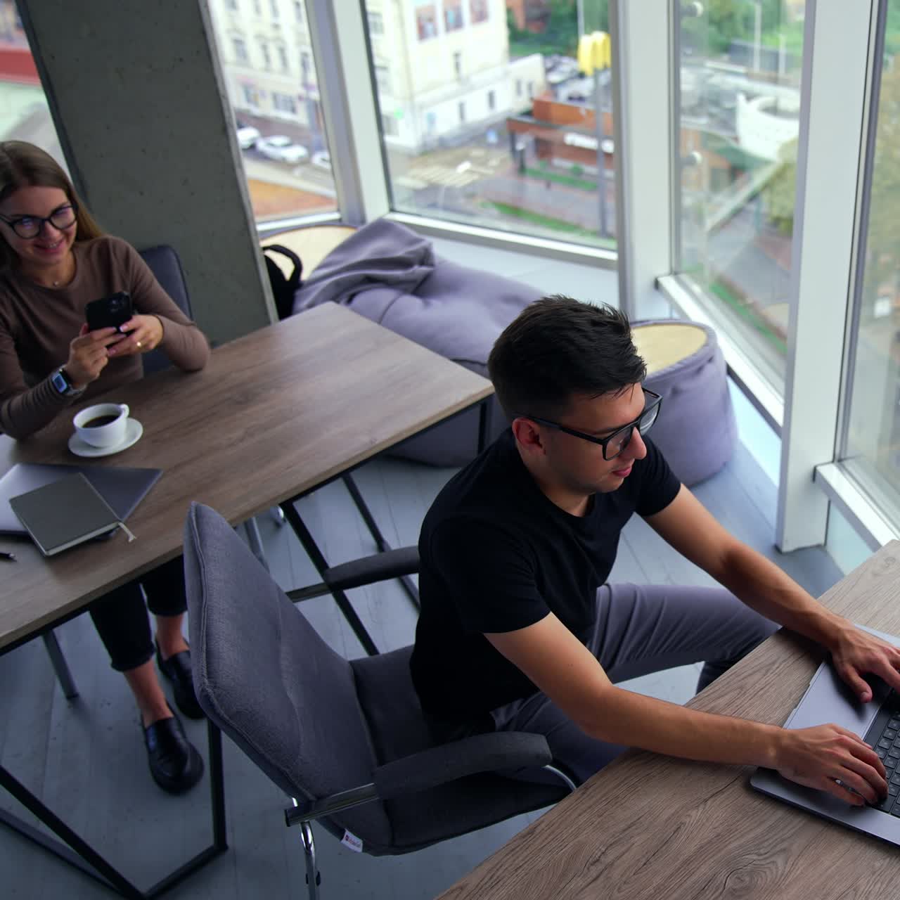 Seorang pria muda beristirahat dari pekerjaan dan memutuskan untuk kembali bekerja. Seorang wanita di meja di belakangnya berbicara di telepon. Kantor dengan jendela panorama dari sudut pandang tinggi. Kota dari atas sebagai latar belakang