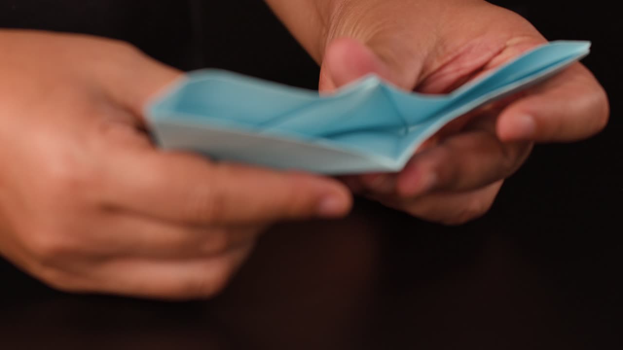 Close-up hands carefully folding blue origami paper boat on dark table, soft studio lighting