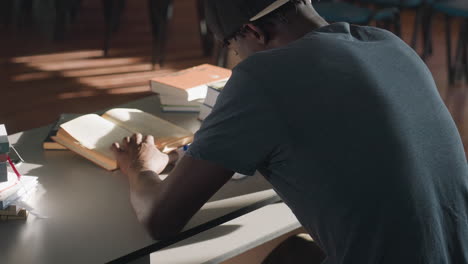 Side view of man sitting at desk in library, reading open book under sunlight while writing notes in notebook, with stacks of books on table and chairs in blurred background