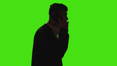 young man wearing wireless headphones answering call on mobile phone against green screen with low key lighting